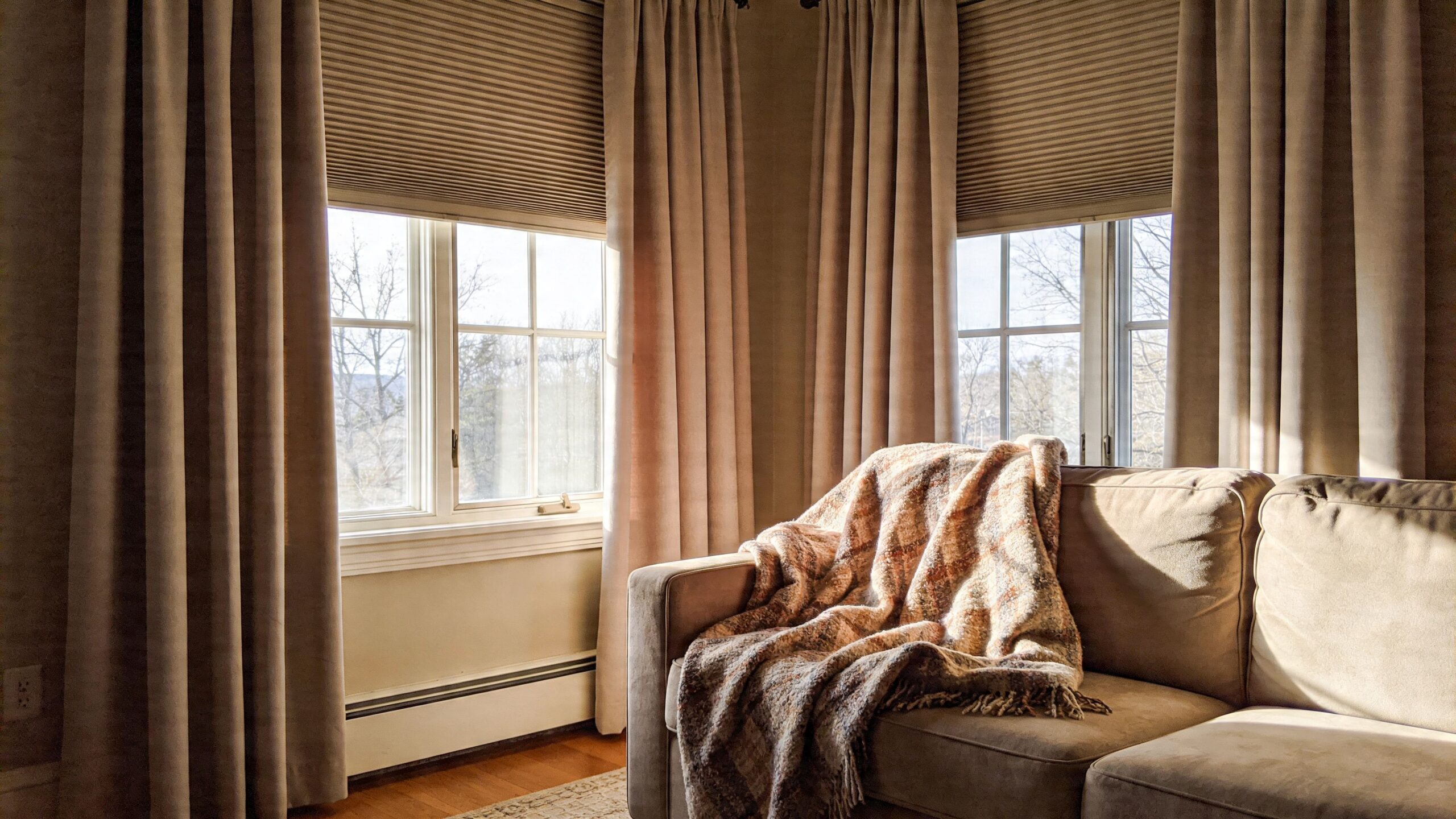 En hyggelig stue med en beige sofa dækket af et tæppe foran solrige vinduer med gardiner.