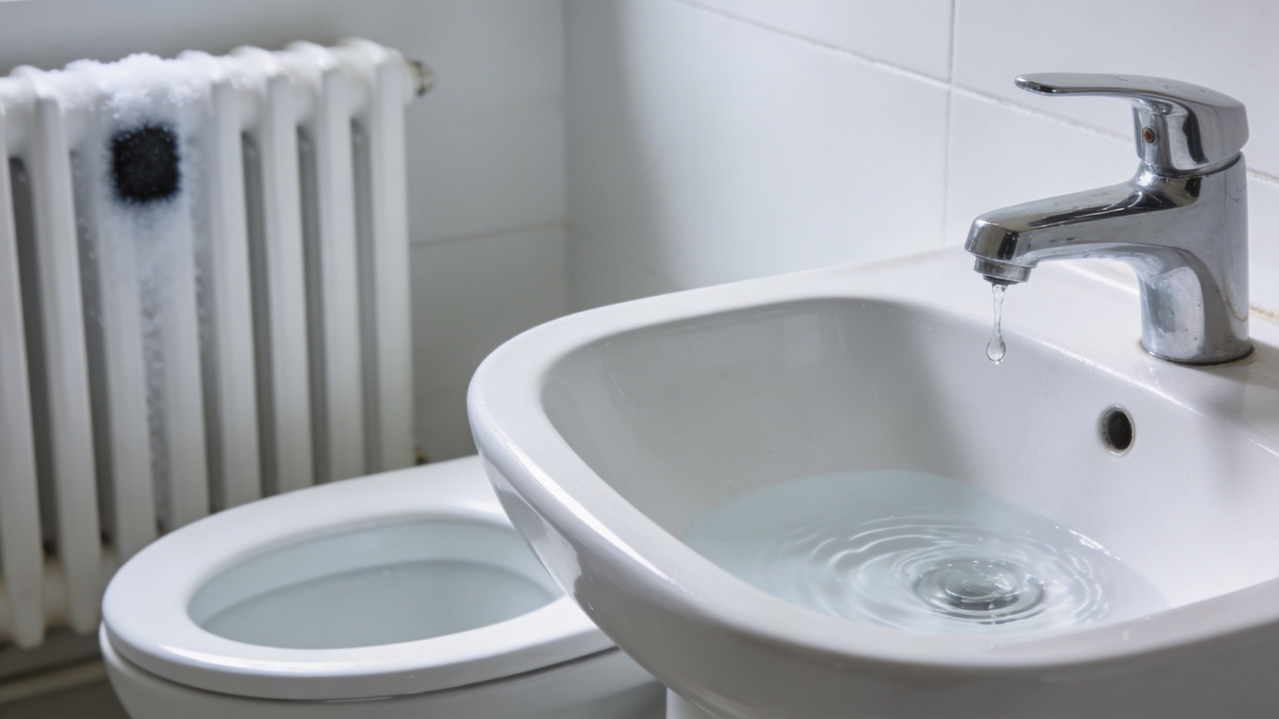 En hvid håndvask med dryppende vandhane og et toilet placeret i et badeværelse med hvide fliser.