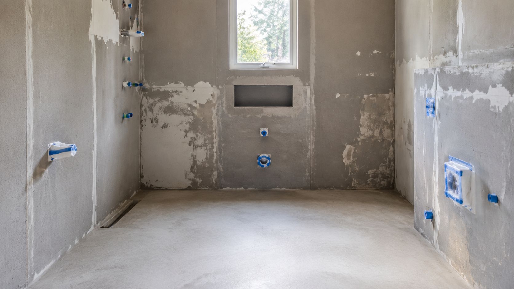 Et ufærdigt badeværelse under renovering med betonvægge, rørføringer og et vindue i midten af bagvæggen.